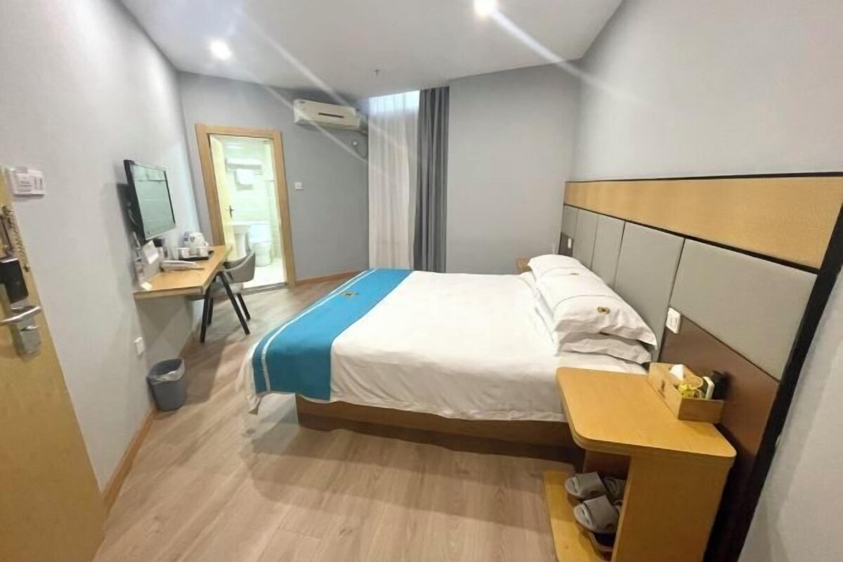 A hotel room with a bed, bedside table, desk, chair, and a small trash bin.