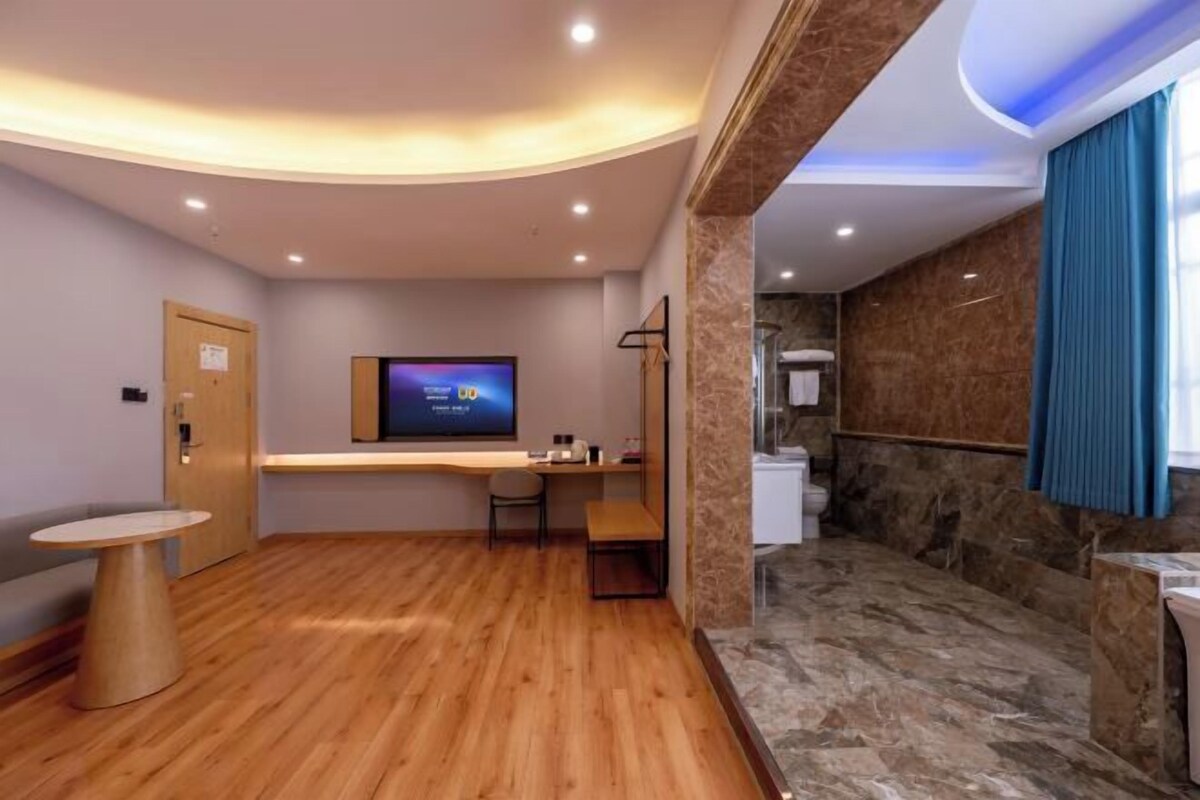 A hotel room with a wooden floor, a flat-screen TV, a desk, a chair, a small table, a bathroom with a marble floor, and a blue curtain.