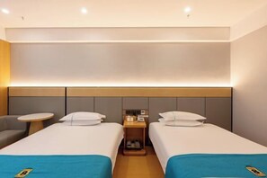 A hotel room with two single beds, a small table, a chair, and a bedside table with a phone and shoe rack.