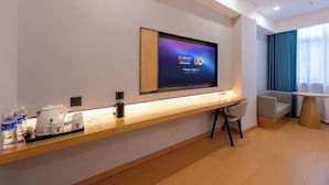 Quarto de hotel moderno com televisão de tela plana na parede, mesa de console de madeira com chaleira e xícaras, pequeno espaço de estar com sofá e poltrona, e uma grande janela com cortinas azuis.