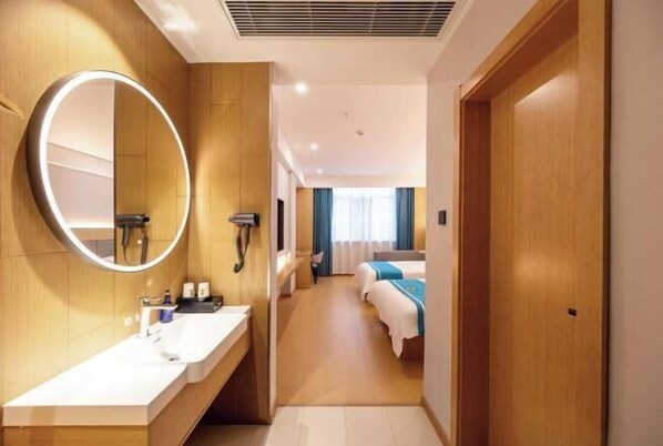 A hotel room with a round mirror, a sink, a shower, and a bed with a blue and white bedspread.