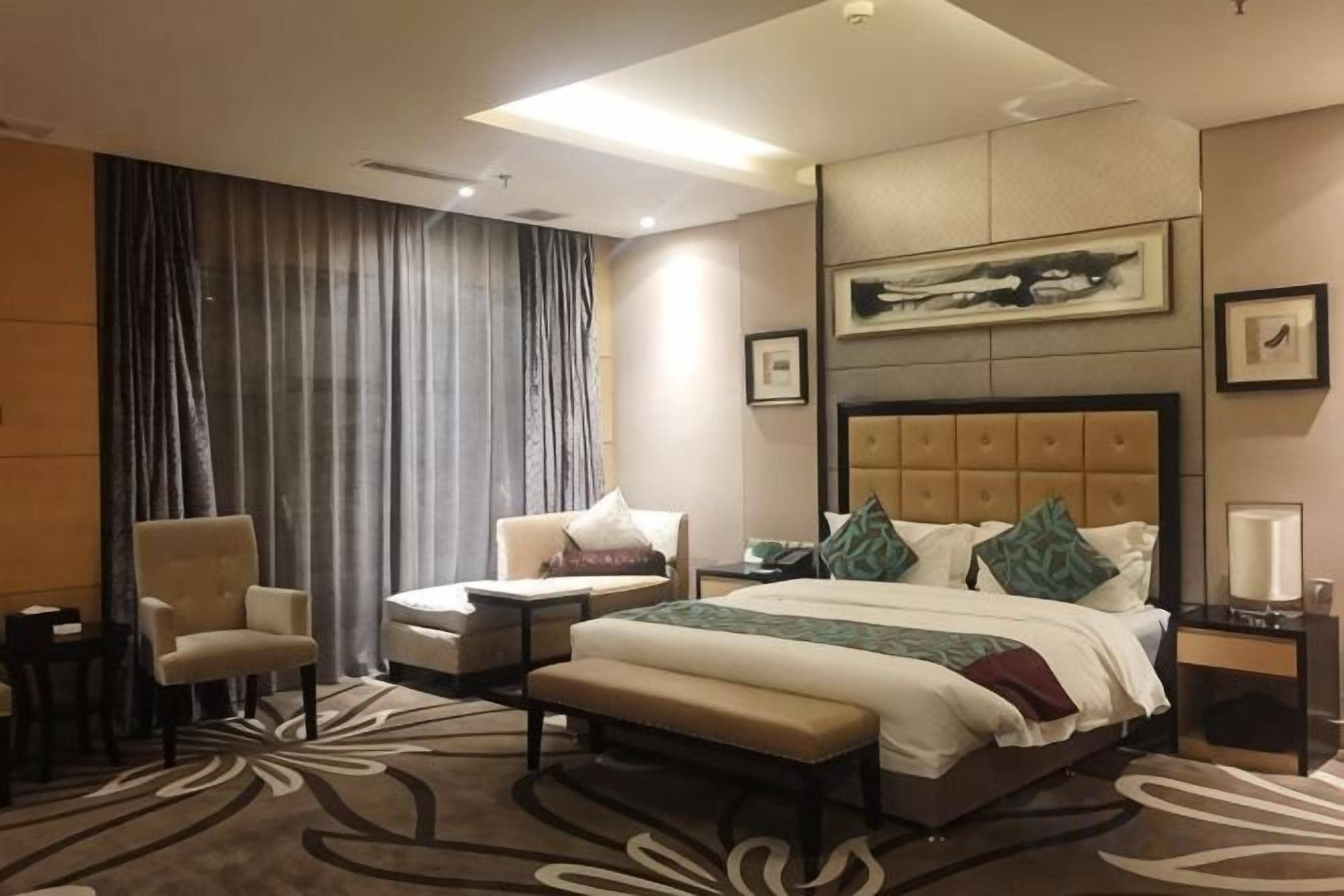 A hotel room with a large bed, two armchairs, a bench, a desk, and a large window with curtains.