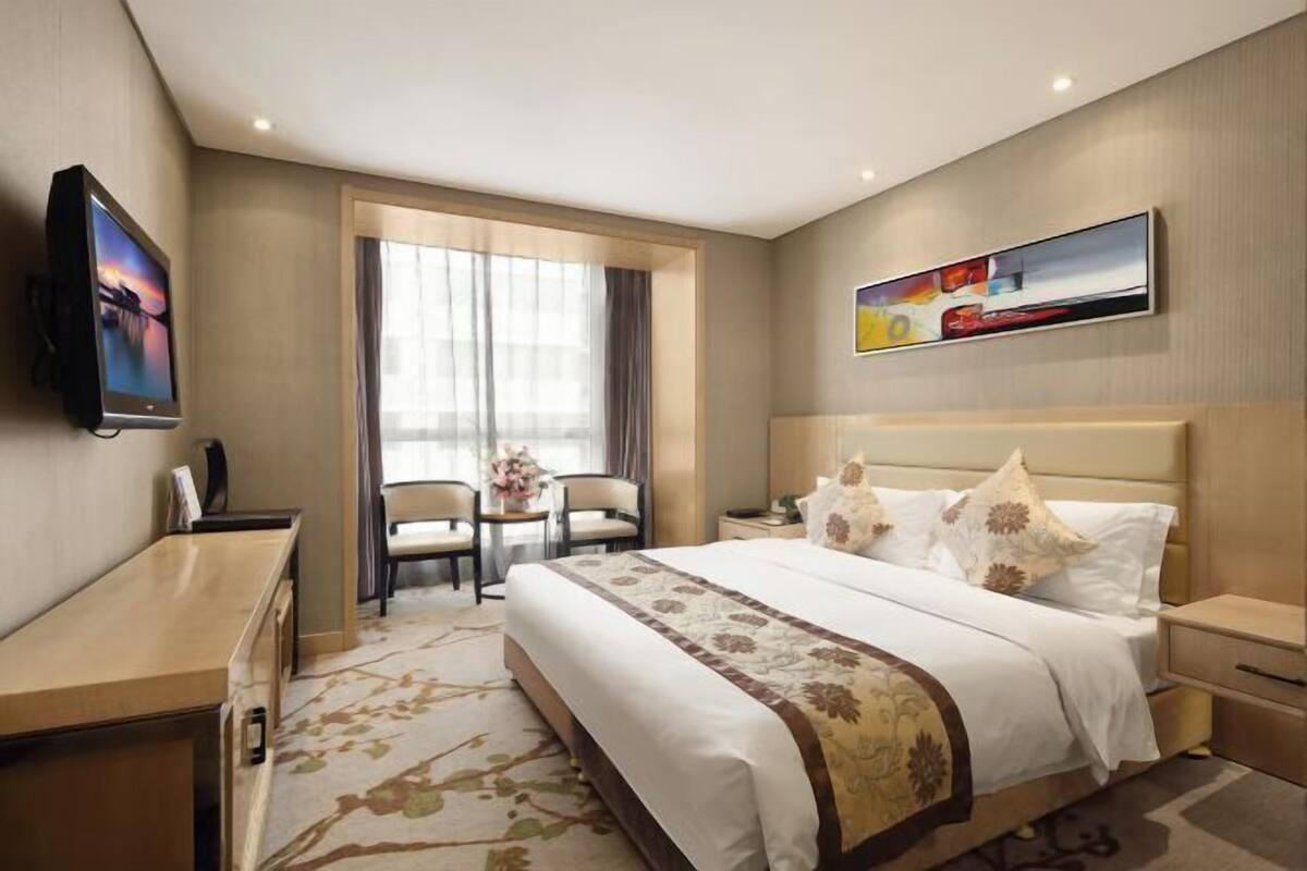 Habitación de hotel con cama, escritorio, televisor, silla, una mesa con flores y un cuadro en la pared.