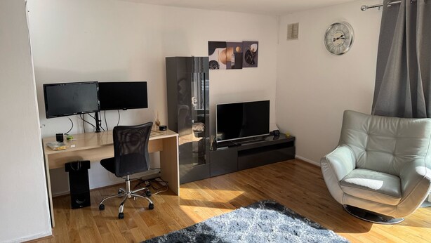 Ein modernes Wohnzimmer mit Holzboden, einem schwarz-weißen Teppich, einem Ledersessel, einem Schreibtisch mit zwei Monitoren, einem Fernseher und einer Wanduhr.