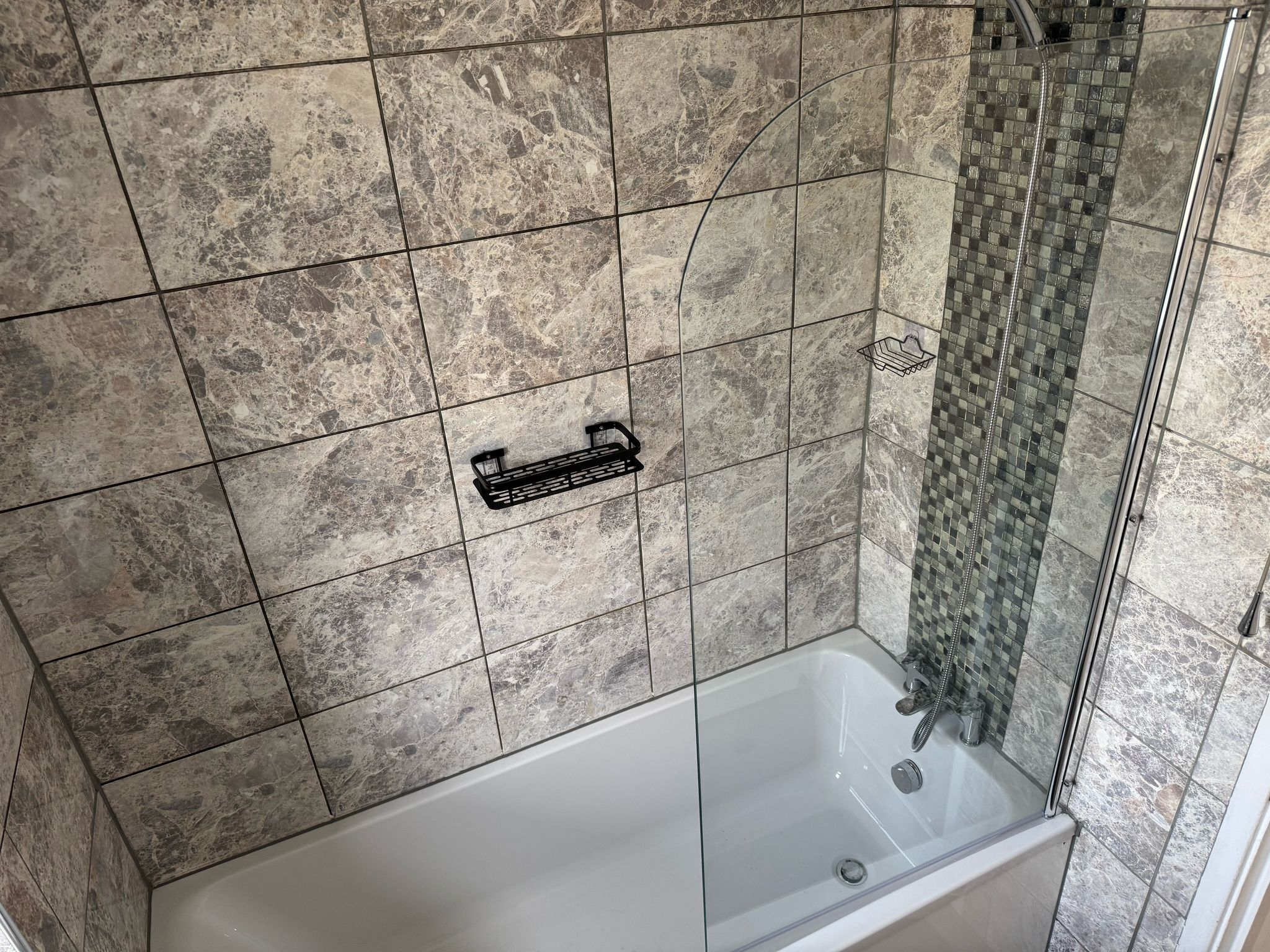 A bathroom with a bathtub, a shower with a glass door, and a tiled wall with a black shelf.