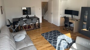 Ein modernes Wohnzimmer mit einem grauen Sofa, einem Esstisch mit Stühlen, einem Fernseher und einer Treppe, die in einen weiteren Raum führt.