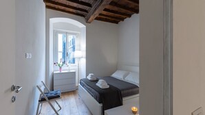 Een slaapkamer met een bed, nachtkastje, lamp en een raam met uitzicht op gebouwen.