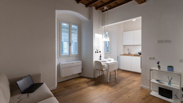 Een moderne, goed verlichte kamer met een houten plafond, een witte radiator, een eettafel met stoelen, een keuken met witte kasten, en een klein plankje met decoratieve objecten.