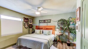A bedroom with a bed, a ceiling fan, a window with blinds, and several potted plants.