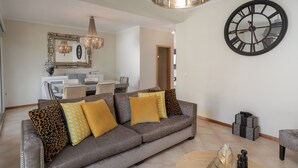 Un salon avec un canapé gris, des coussins jaunes et à motifs léopard, une table basse ronde et une grande horloge murale.