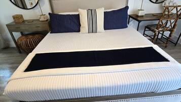 Uma cama bem arrumada com cabeceira, duas mesas de cabeceira com abajures, uma cadeira e um painel de parede texturizado.