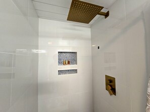 Un baño moderno con cabina de ducha de vidrio, estante empotrado con objetos decorativos y panel de control de ducha dorado.