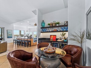 Un vaste espace de vie comprenant un salon avec une table basse en verre, des fauteuils en cuir, une cuisine avec un bar et une grande fenêtre offrant une vue panoramique.