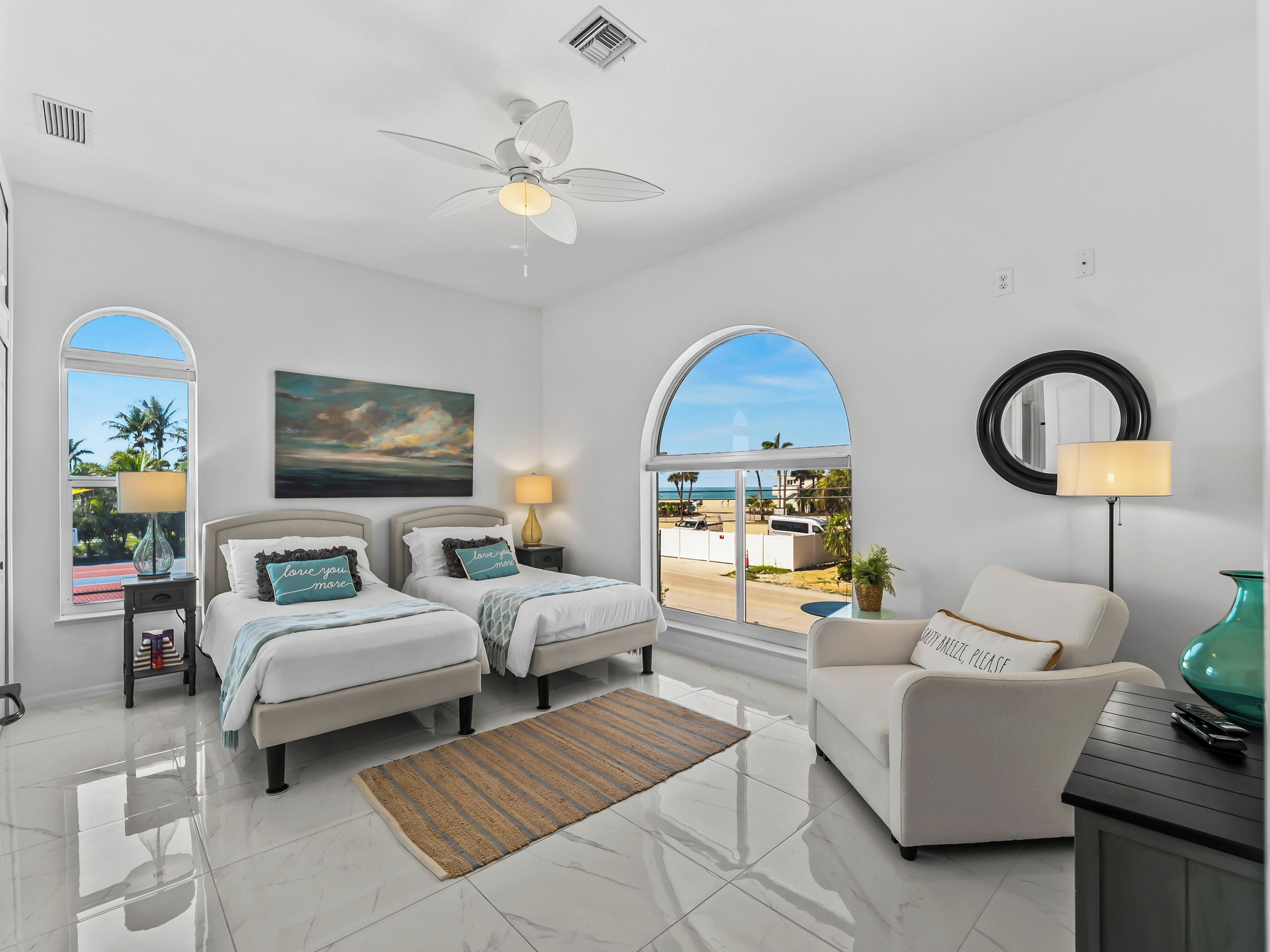 A hotel room with two beds, a ceiling fan, a large window with a view, a round mirror, and a side table with a lamp.