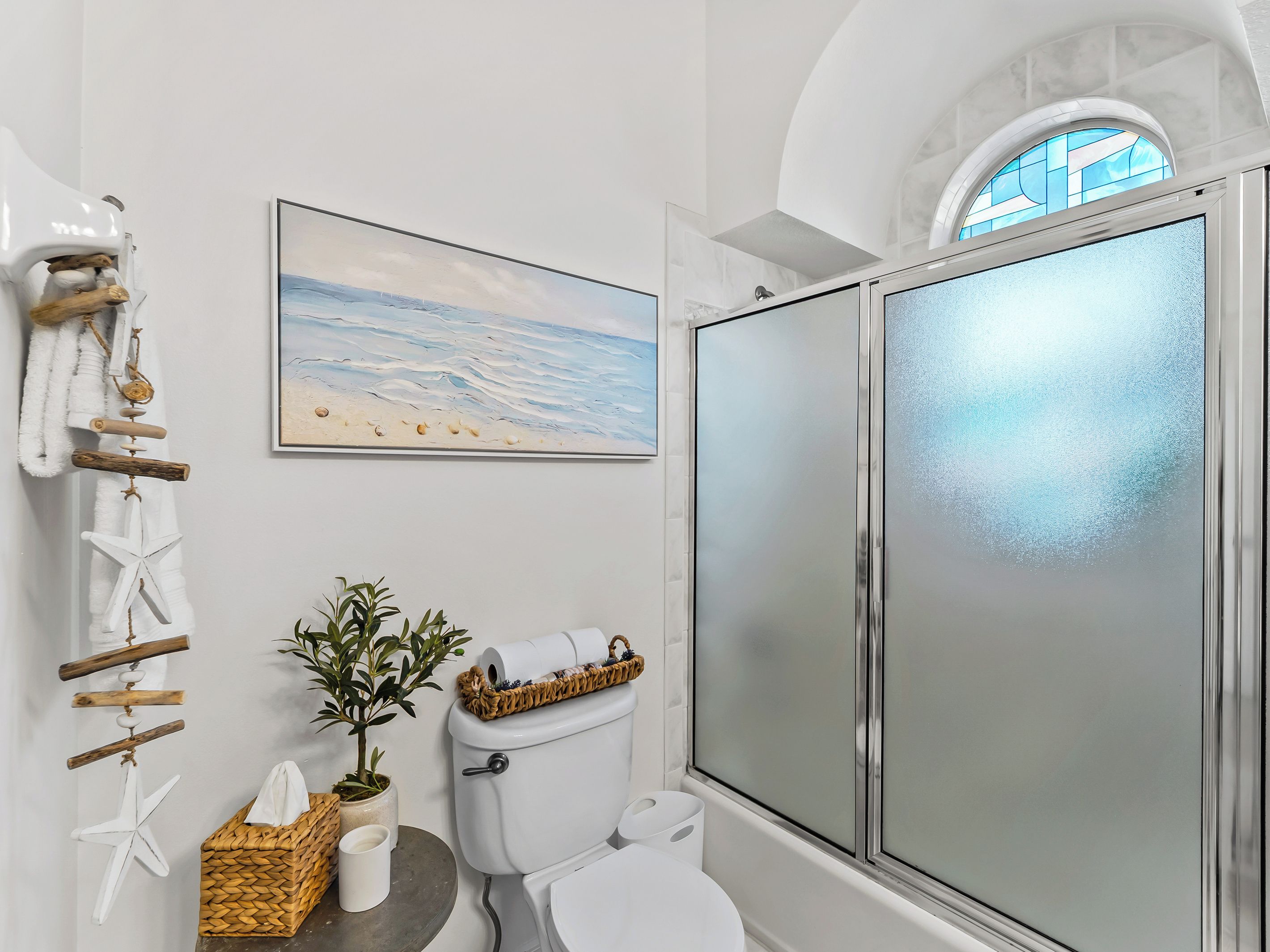 Un baño con inodoro, un bidé, cabina de ducha de vidrio, un cuadro de una playa enmarco y una decoración con forma de estrella de mar.