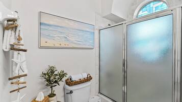 Un baño con inodoro, un bidé, cabina de ducha de vidrio, un cuadro de una playa enmarco y una decoración con forma de estrella de mar.