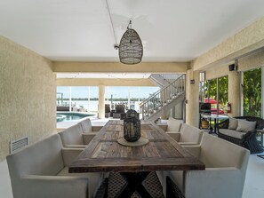 Un área de comedor al aire libre con una mesa de madera, sillas acolchadas, un colgador de luz y vistas a una piscina y una escalera que conduce a un nivel superior.
