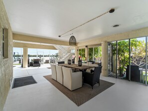 A spacious outdoor dining area with a wooden table and chairs, a staircase leading to an upper level, and large windows offering views of the surrounding landscape. - Casa Bahia-Luxury Bayfront Pool Home with Dock (Englewood)