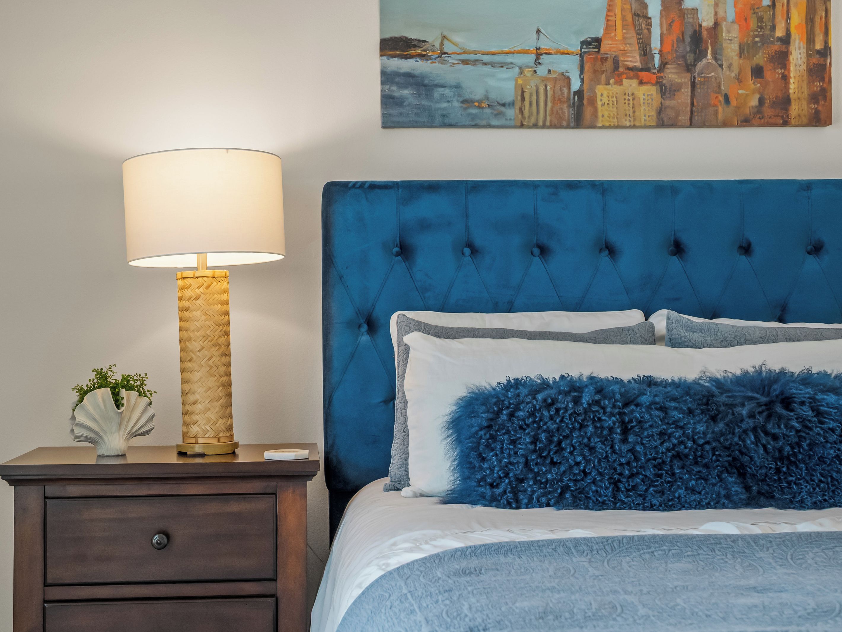 A bedroom with a tufted headboard, a bedside lamp, a nightstand, and a painting of a cityscape.