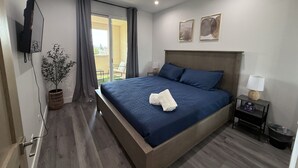 Une chambre d’hôtel moderne dotée d’un grand lit, d’un téléviseur fixé au mur, d’un balcon avec des rideaux et d’une petite table avec une lampe.