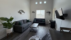Un salon moderne avec un canapé gris, une table basse et une table à manger. Il y a trois petites fenêtres équipées de stores, un téléviseur fixé au mur et une plante en pot.