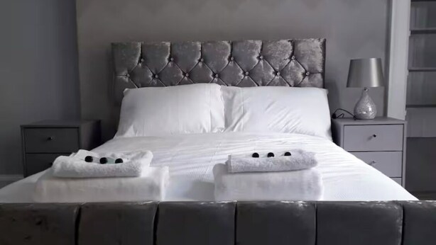 Una cama con cabecera capitonada, ropa de cama blanca y dos toallas sobre la cama.