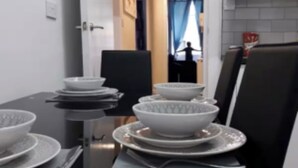 Un comedor con una mesa negra dispuesta para cuatro personas, platos y cuencos blancos, y un vaso. Hay dos sillas, una negra y una blanca. Un acceso da a una zona de cocina con salpicadero de azulejos y una ventana con cortinas.