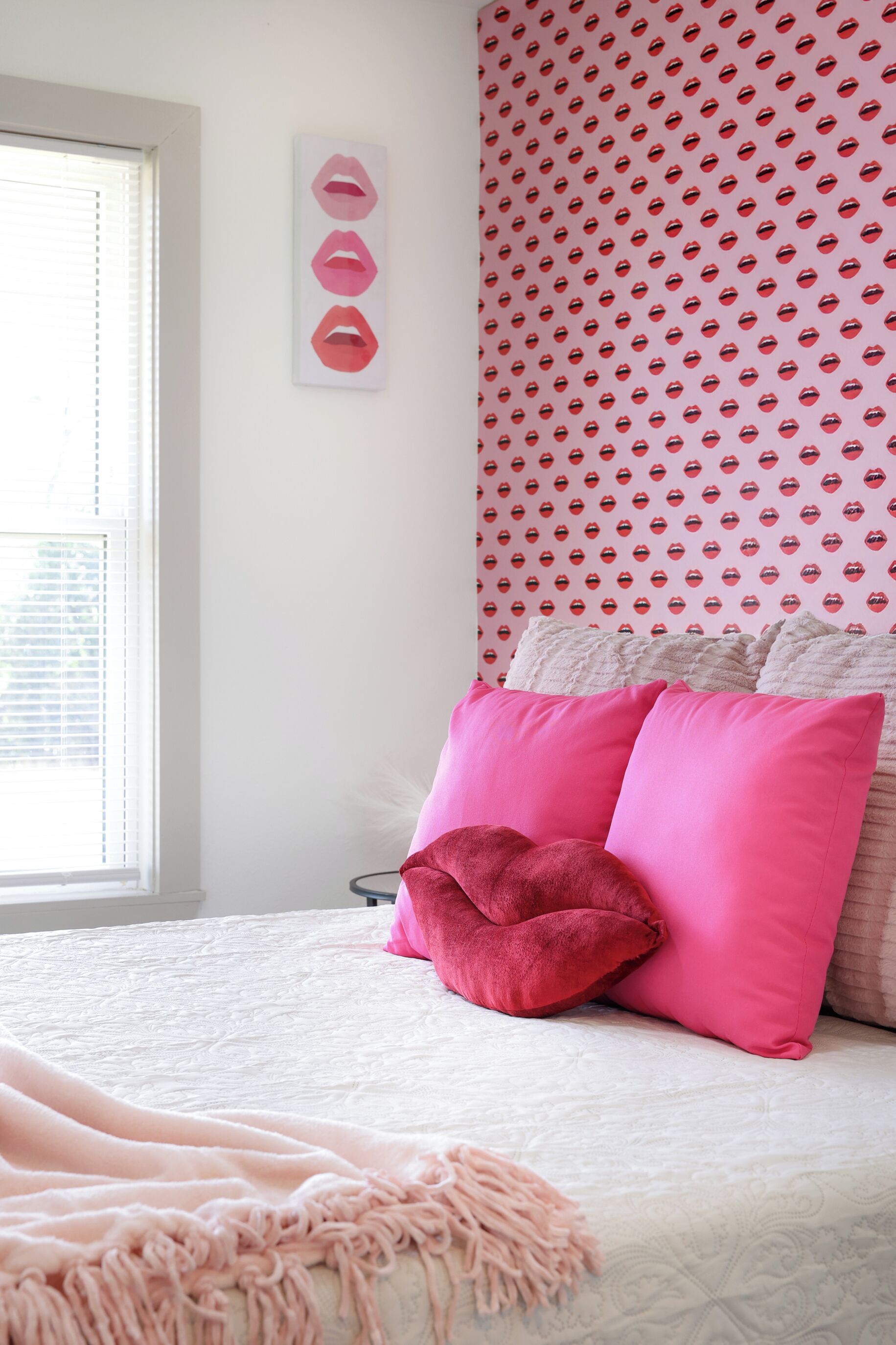 Une chambre à coucher avec un lit, des coussins roses, un mur à motifs et une fenêtre avec des stores.