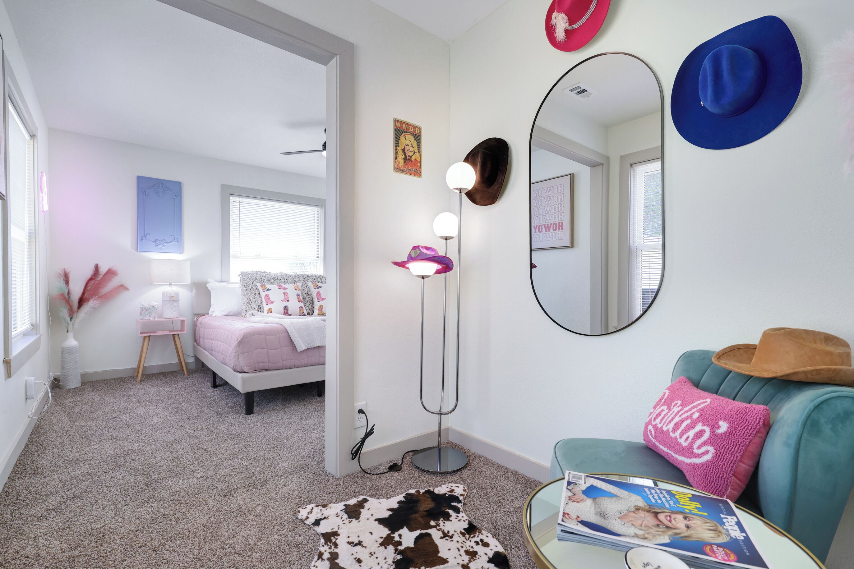Une chambre à coucher moderne avec un lit, une lampe sur pied, un miroir et un canapé bleu agrémenté d’un chapeau et d’un coussin rose.
