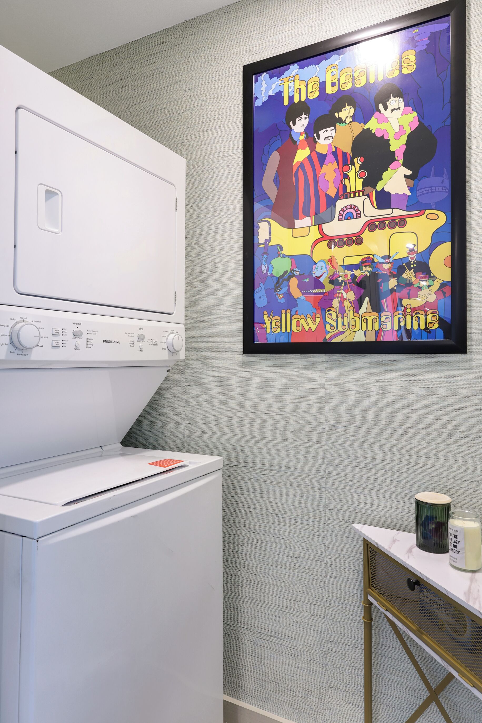A laundry area with a white washing machine and dryer, a wall-mounted poster of The Beatles' 'Yellow Submarine', and a small table with a candle and a container.