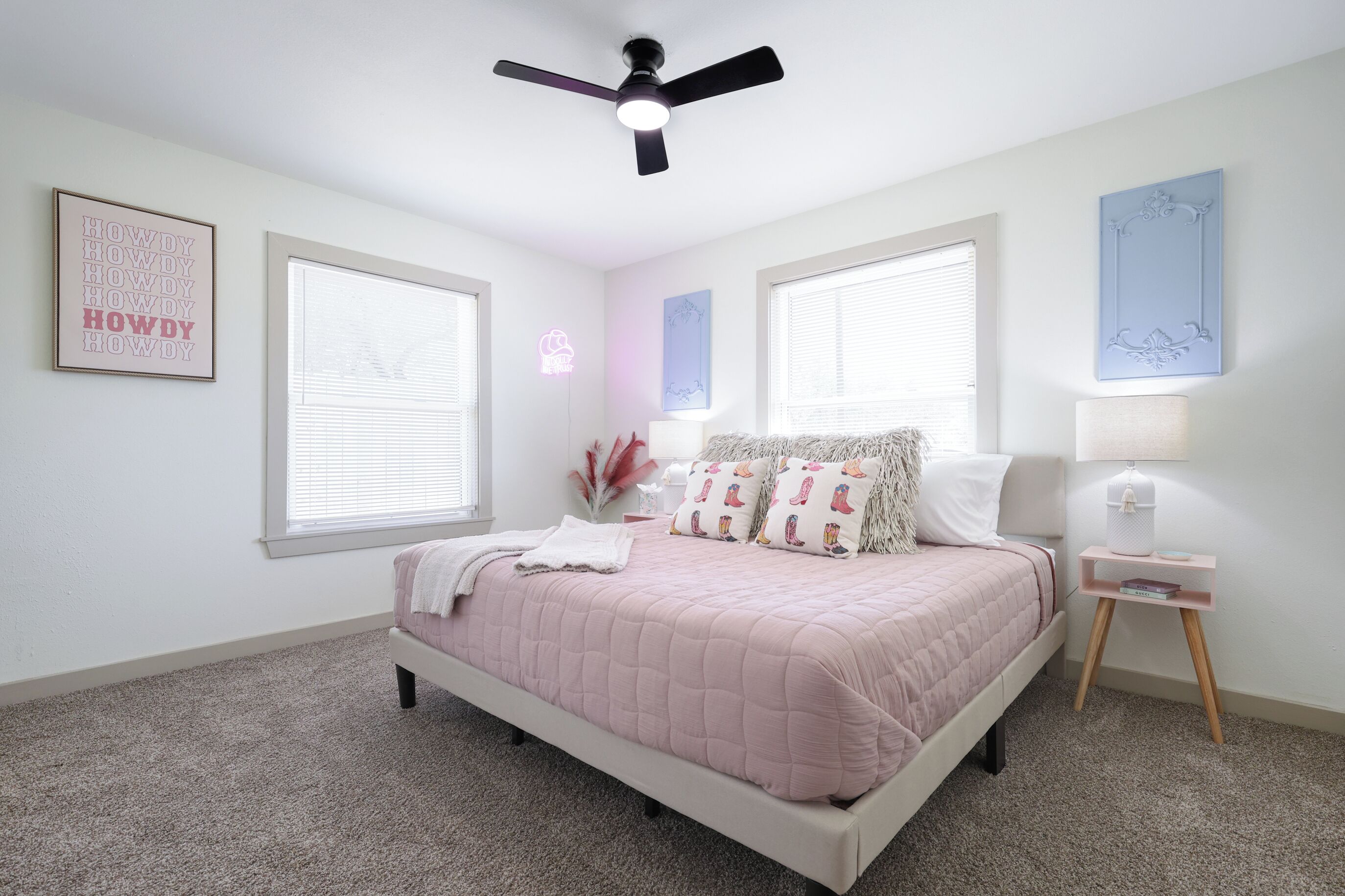 Un dormitorio con cama, ventilador de techo, dos ventanas, mesita de noche con lámpara y decoración en la pared.