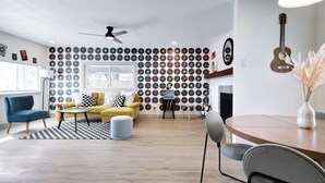 Een moderne woonkamer met een wand vol vinylplaten, een gele bank, een blauwe fauteuil en een eettafel met stoelen.