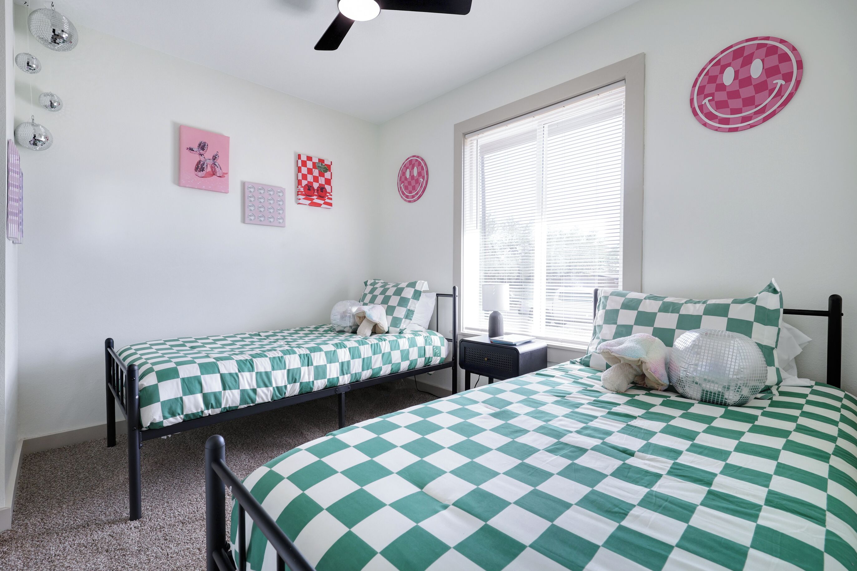 Un dormitorio con dos camas individuales, una ventana con persianas, y decoraciones en la pared incluyendo una carita sonriente y un diseño a cuadros.