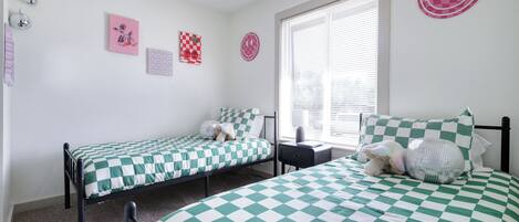Un dormitorio con dos camas individuales, una ventana con persianas, y decoraciones en la pared incluyendo una carita sonriente y un diseño a cuadros.