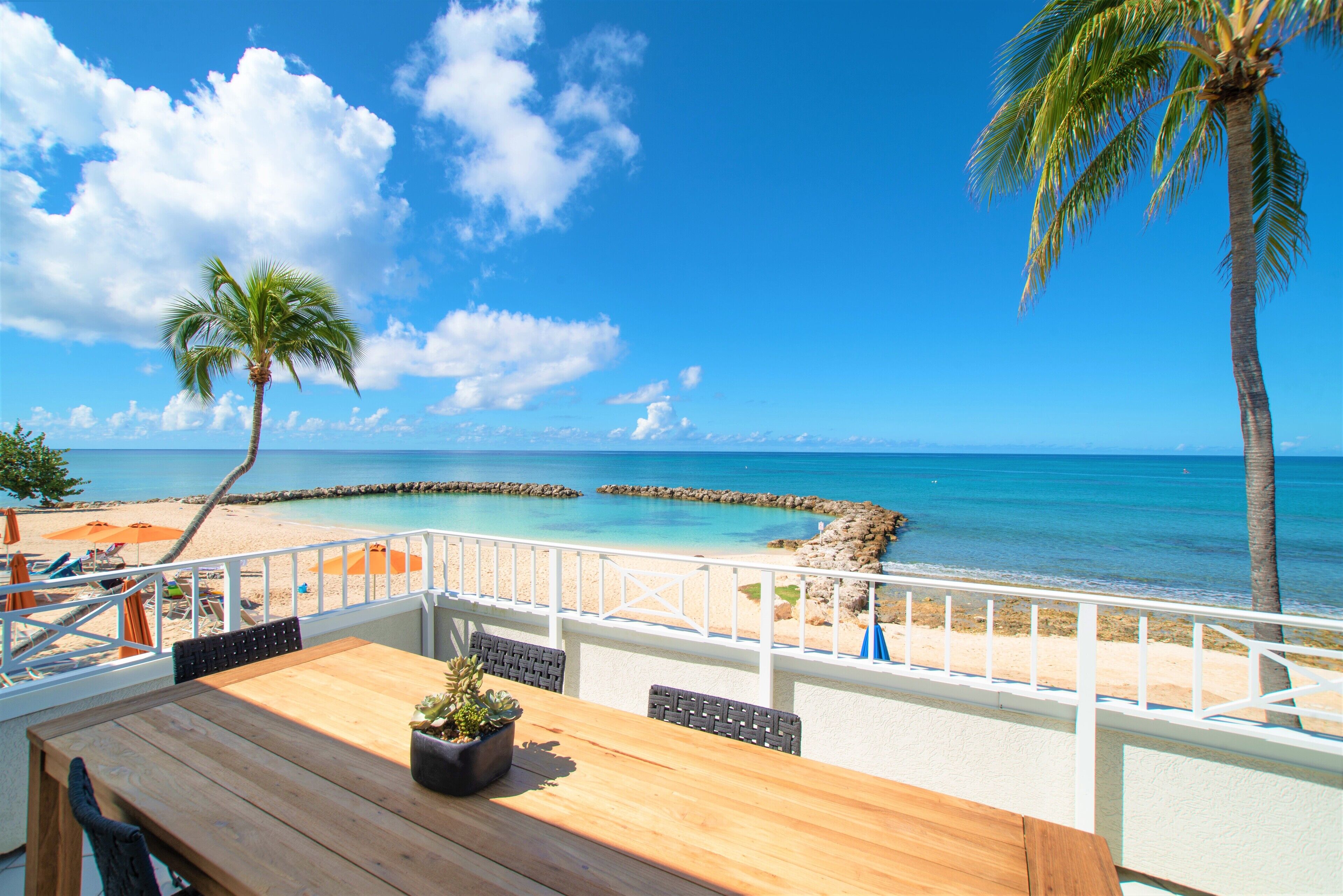 A wooden deck with a table and chairs, overlooking a beach with a palm tree and a clear blue sky.
