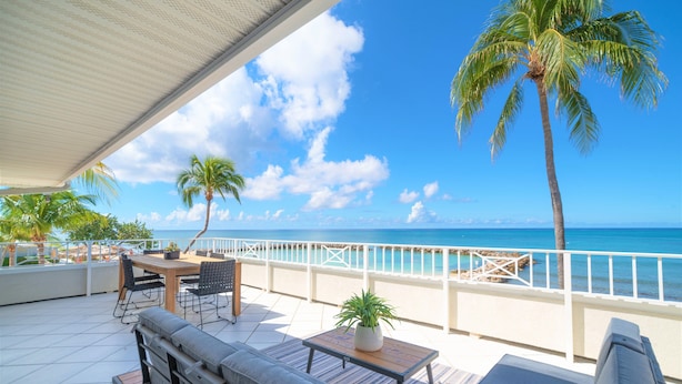 A terrace with a table, chairs, and a sofa overlooking a beach with palm trees and the ocean. - Cayman Luxury Rentals at Sunset Cove (Seven Mile Beach)