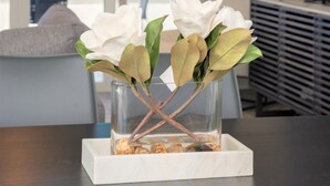 A modern living room with a large window, a flat-screen TV, and a vase with white flowers on a table. - Cayman Luxury Rentals at Sunset Cove (Seven Mile Beach)