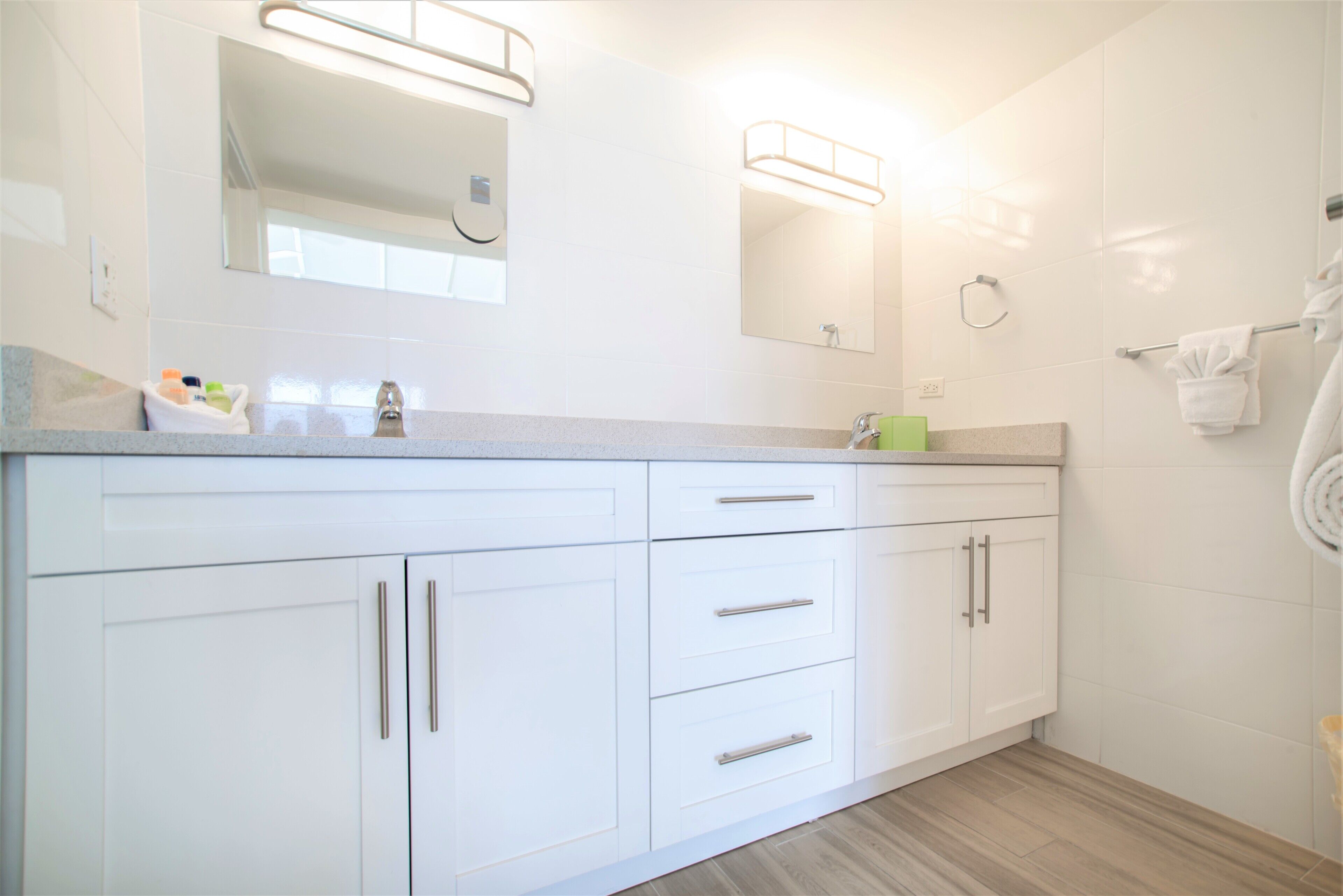Une salle de bain moderne avec des meubles blancs, un plan de toilette où sont posés les articles de toilette, un miroir et un porte-serviettes.