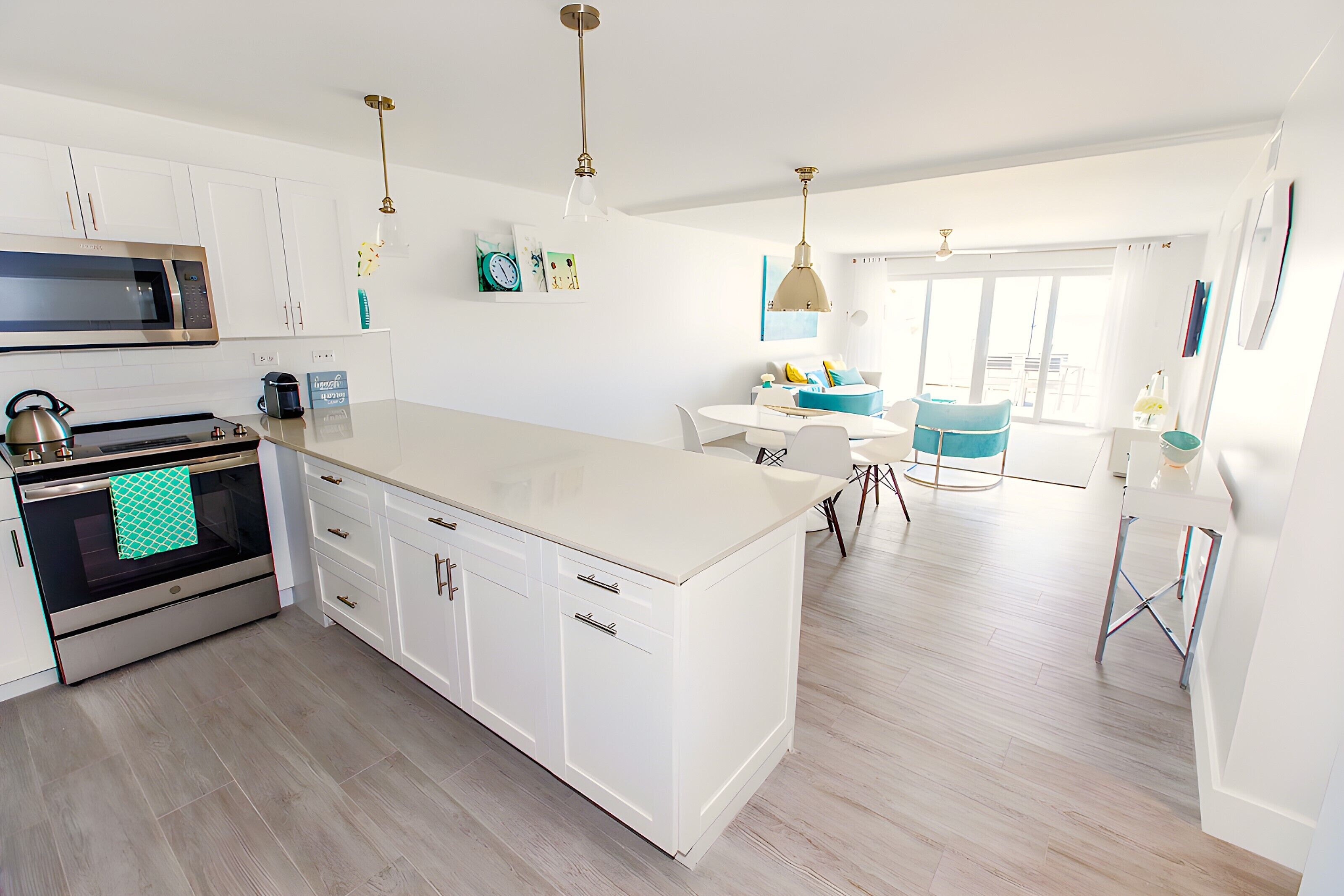 A modern kitchen with white cabinets, stainless steel appliances, and a large island. There is a dining area with a blue chair and a table.