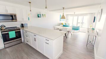 A modern kitchen with white cabinets, stainless steel appliances, and a large island. There is a dining area with a blue chair and a table.