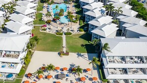 An aerial view of a resort with multiple buildings, a central pool area, and a beach with sun loungers and umbrellas. - Cayman Luxury Rentals at Sunset Cove (Seven Mile Beach)