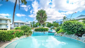 A resort pool area with a clear blue pool, palm trees, and lush greenery. - Cayman Luxury Rentals at Sunset Cove (Seven Mile Beach)
