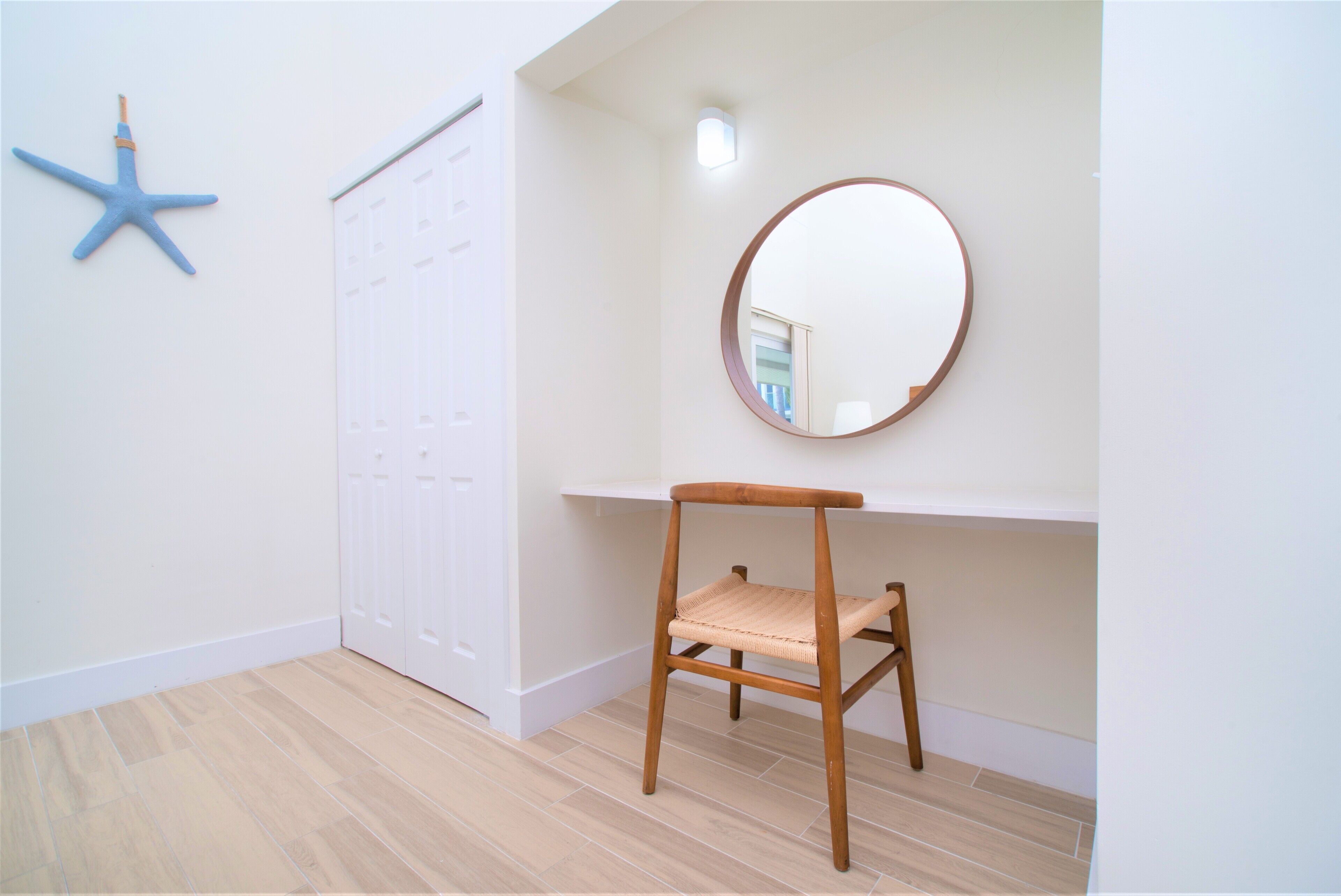 Una habitación con una silla de madera, un espejo redondo, una decoración de estrella de mar y una puerta blanca.