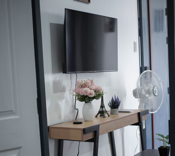Una sala de estar moderna con una televisión de pantalla plana montada en la pared, una mesita de madera con un jarrón de flores, una pequeña réplica de la Torre Eiffel y un ventilador de pie.