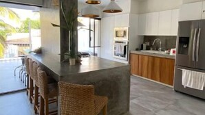Une cuisine moderne dotée d’un îlot central, de tabourets de bar, de luminaires suspendus et d’un réfrigérateur en acier inoxydable.