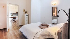 A bedroom with a bed, bedside lamp, and a mirror. There is a door leading to a bathroom and a kitchenette. - Apartments Florence I Portici (Firenze)