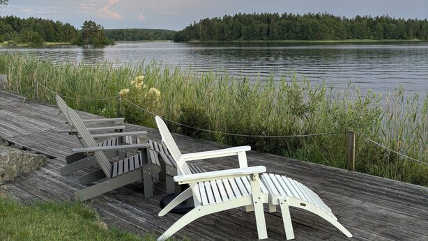 A wooden deck with two Adirondack chairs facing a lake, surrounded by grass and reeds.