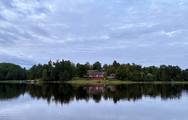 Une propriété au bord du lac, avec une maison rouge, entourée d’une forêt dense et d’une eau calme qui reflète le ciel.