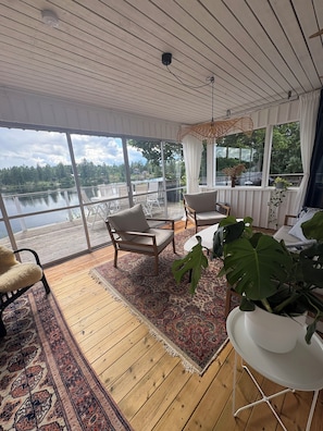 Ett rum med ett stort fönster med utsikt över en sjö, ett trägolv och en sittgrupp med stolar och ett bord.