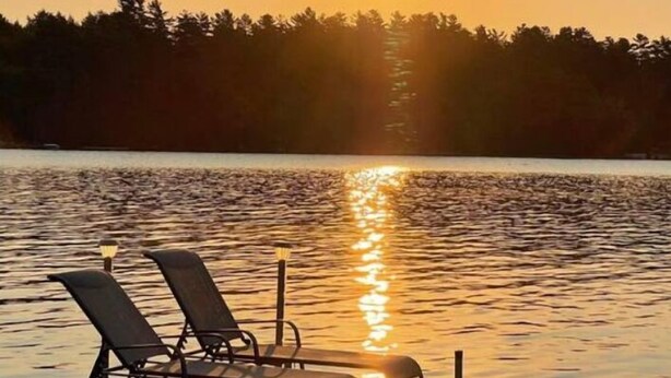 A wooden dock with two lounge chairs facing a calm lake at sunset.